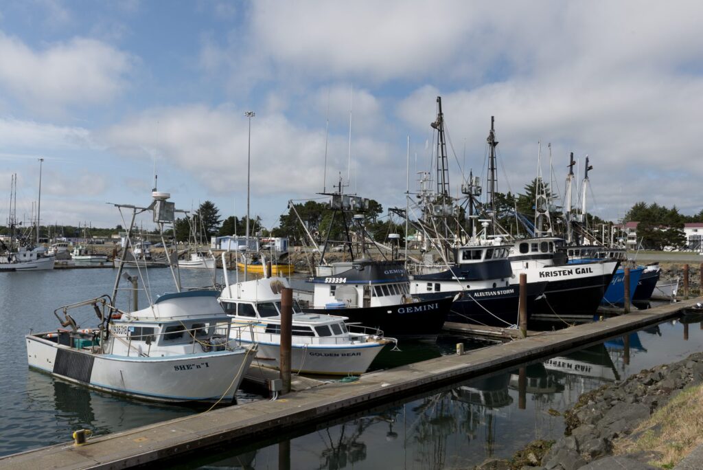 Commercial fishing boats with tall rigging tied up along a wooden dock in Crescent City's harbor under a partly cloudy sky.