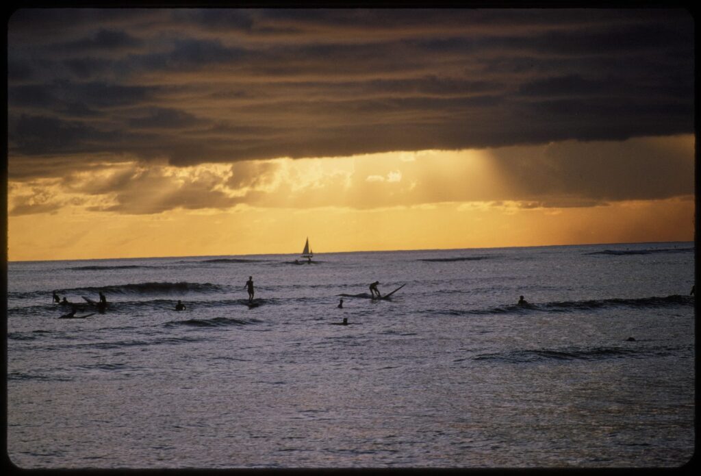 Silhouetted surfers sit and ride small waves under a dark band of storm clouds as golden light breaks over the horizon and a sailboat passes in the distance.