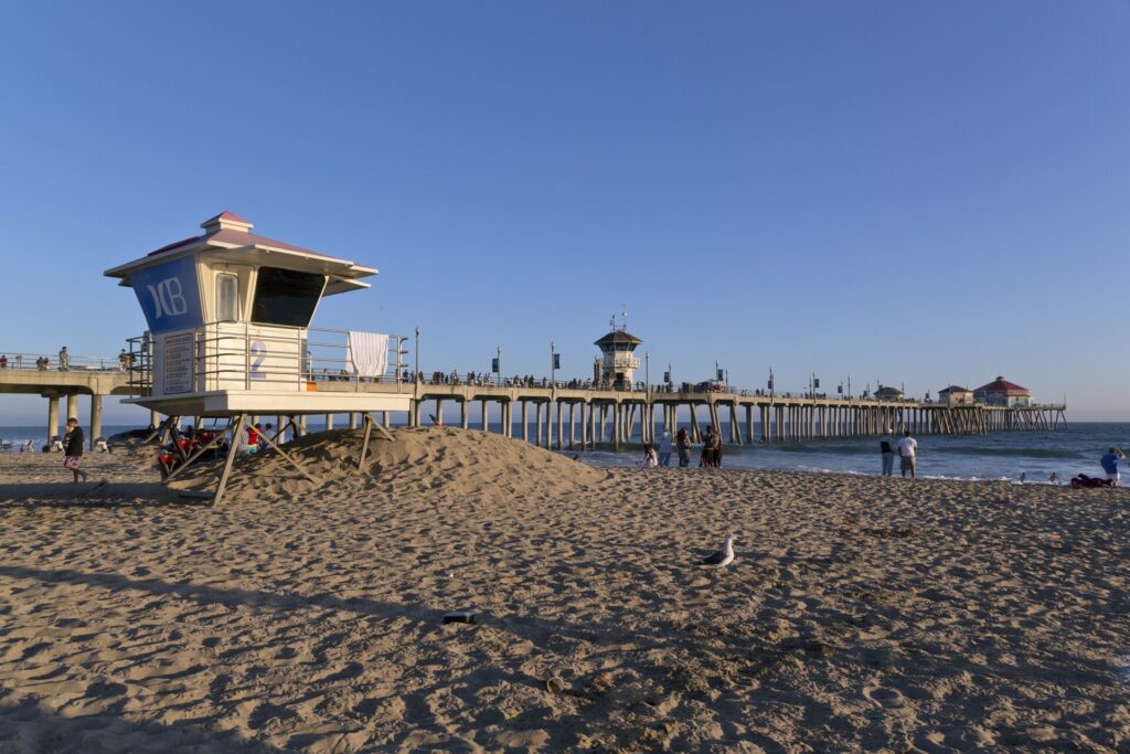 A blue-and-white lifeguard tower on a sandy mound in the foreground with the long concrete Huntington Beach Pier stretching out to sea behind it under a clear evening sky.