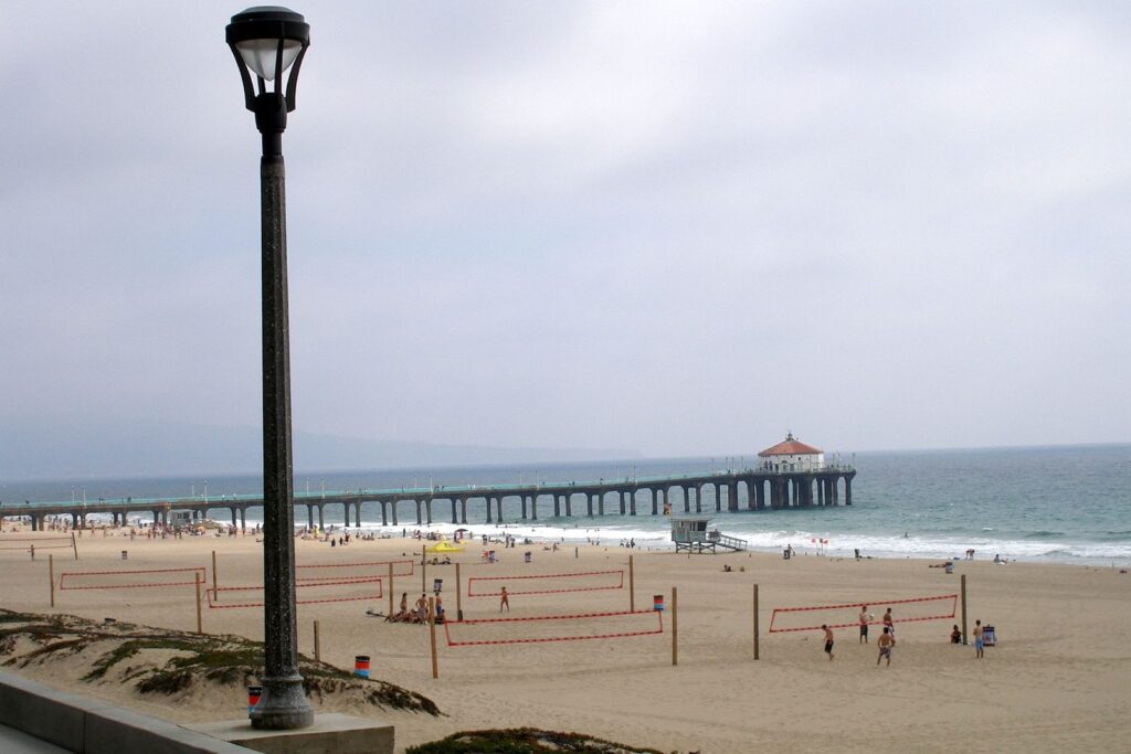 A wide beach with volleyball nets and a lifeguard tower stretches toward the Manhattan Beach Pier and its red-roofed roundhouse under a gray marine-layer sky, with an old-fashioned lamppost in the foreground.