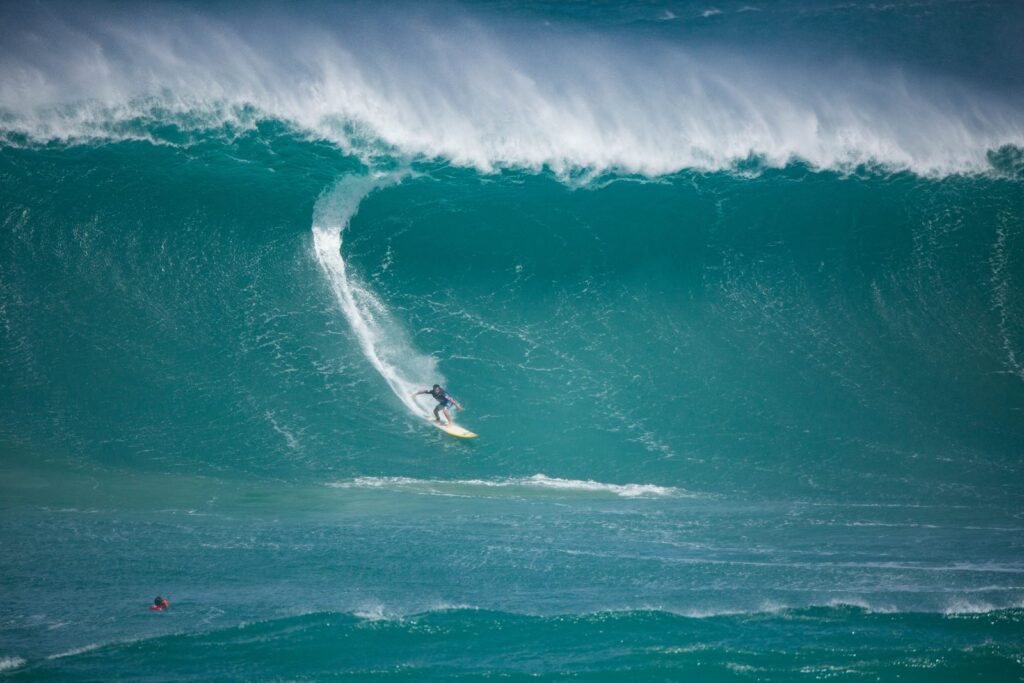 A lone surfer carves down the face of a giant green wave at Waimea Bay as the lip feathers white overhead, with a water-safety swimmer bobbing in the foreground.